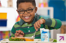 Young African American boy sitting at lunch table in front of food tray and smiling.